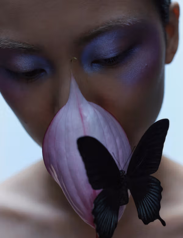A person with violet eye makeup behind a purple flower and a black butterfly.