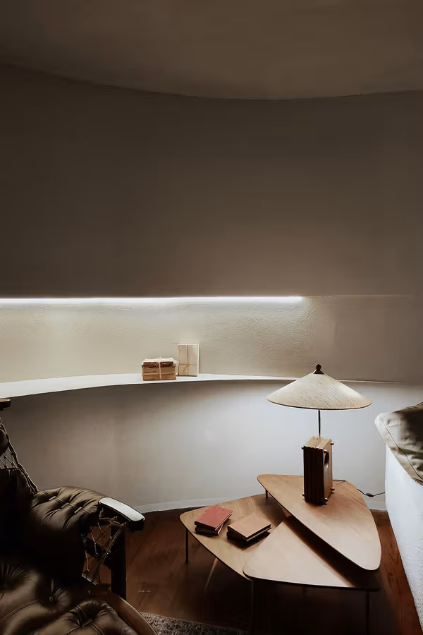 A dim sitting room with a curved illuminated shelf and a lamp on a low table.
